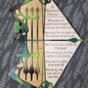 May include: A wooden sign shaped like a bow and arrow with a green and brown paint design. The sign has a poem on it that reads: "A Hunter's Prayer. We pray our sights be straight and our aim be true. We pray for no pain to the game we pursue. We thank you LORD for this land. We thank you for the sights in our stands. We pray for safety one and all. We pray we may return next Fall."