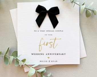 Puede incluir: Una tarjeta de felicitación cuadrada de color blanco con un lazo de tela negro en la parte superior. El texto dice: TO A VERY SPECIAL COUPLE ON YOUR first WEDDING ANNIVERSARY. Debajo hay un pequeño corazón dorado y los nombres MILLIE & RYAN.