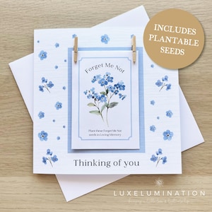 May include: A white card featuring blue forget-me-not flower illustrations and the text "Forget Me Not" and "Thinking of you." Includes plantable seeds. The card is held by two small wooden clips.