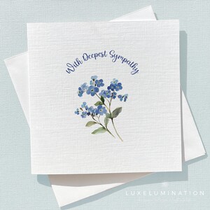 May include: A white sympathy card with the words "With Deepest Sympathy" in blue script. The card features a watercolour illustration of blue forget-me-not flowers. The card is placed on top of a white envelope.