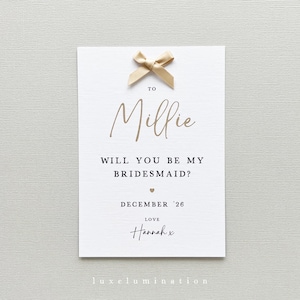 May include: A white card with gold text and a gold bow. The card reads "TO Millie, WILL YOU BE MY BRIDESMAID?" with the date "DECEMBER '26" and the name "Hannah x" at the bottom. The card is a wedding invitation.