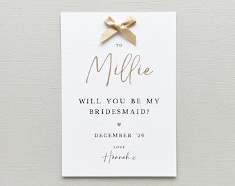 May include: A white card with gold text and a gold bow. The card reads "TO Millie, WILL YOU BE MY BRIDESMAID?" with the date "DECEMBER '26" and the name "Hannah x" at the bottom. The card is a wedding invitation.