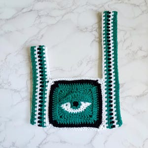May include: A crochet crop top featuring a square central panel with a stylized eye motif in teal, white, and black. The garment includes two wide shoulder straps with a repeating vertical striped pattern in teal, white, and black yarn.