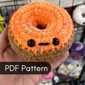 May include: A crocheted orange donut with brown frosting and colorful sprinkles. The donut has googly eyes and a black mouth. The text "PDF Pattern" is visible at the bottom of the image.