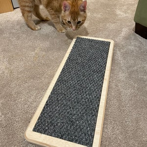 May include: A rectangular cat scratching board featuring a light-colored wooden frame and a textured dark gray carpet insert. The scratching surface is flat and elongated, designed for feline use, with rounded corners on the wooden border.