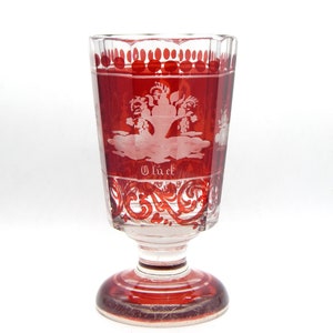 May include: A red glass goblet with a white etched design of a tree and the word "Glück" on the bottom. The goblet has a wide base and a flared top.