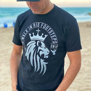 May include: A black t-shirt with a white graphic design of a lion's head with a crown and the text "Walk in His Footsteps Est. 2018".