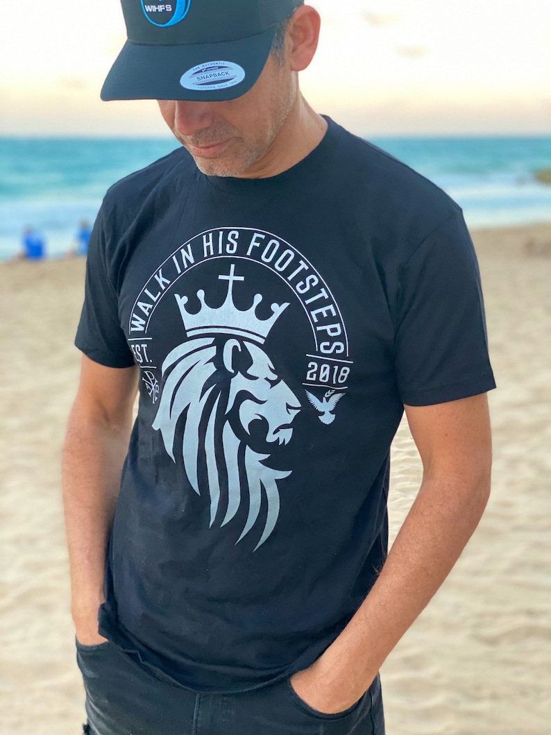 May include: A black t-shirt with a white graphic design of a lion's head with a crown and the text "Walk in His Footsteps Est. 2018".