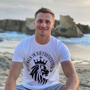 May include: A white t-shirt with a black lion graphic and the text "Walk in His Footsteps" in a circle around the lion. The lion is wearing a crown and there is a cross above the crown. The text "Est. 2018" is below the lion.