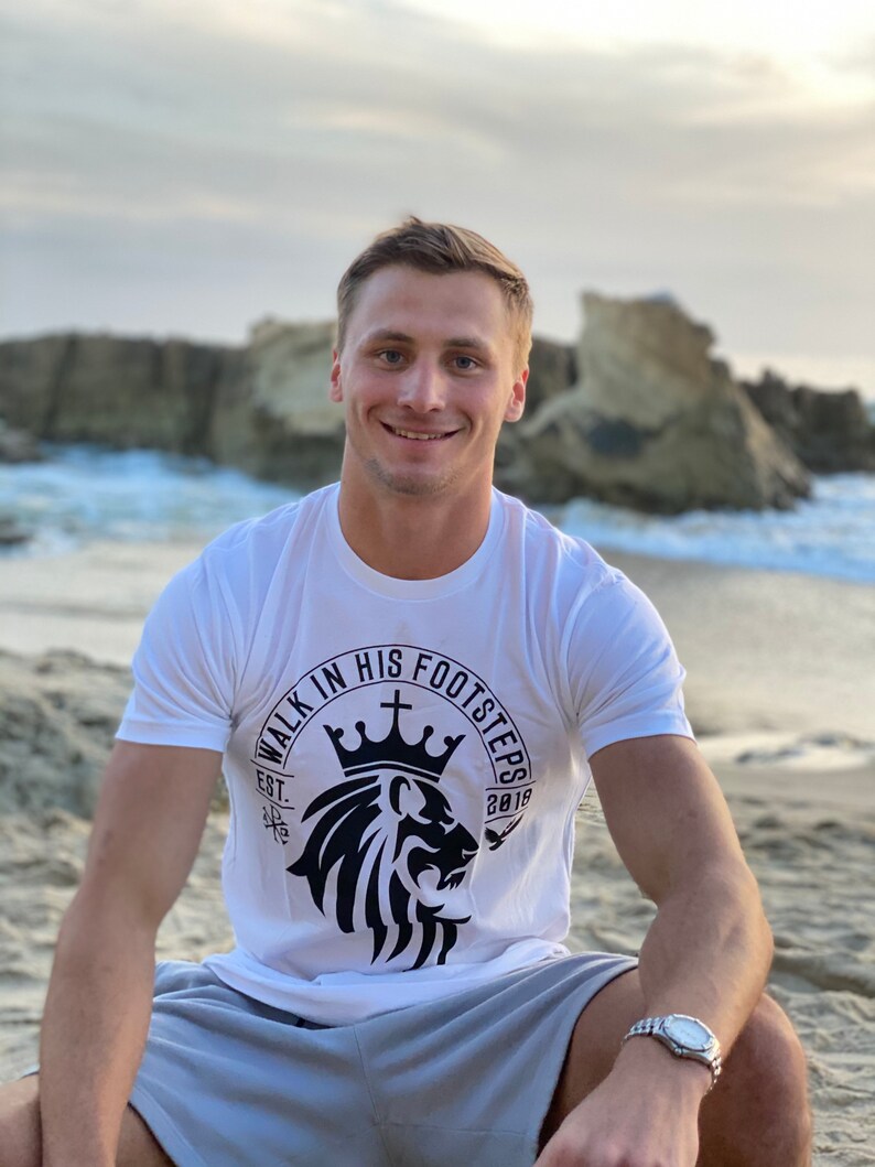 May include: A white t-shirt with a black lion graphic and the text "Walk in His Footsteps" in a circle around the lion. The lion is wearing a crown and there is a cross above the crown. The text "Est. 2018" is below the lion.