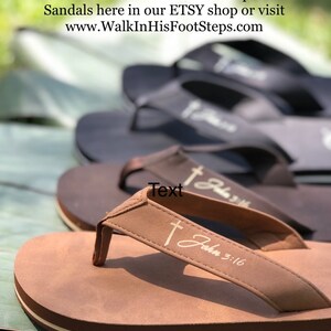 Op de afbeelding: Close-up van een bruine leren sandaal met een wit kruis en de tekst "Johannes 3:16" op de band. De sandaal heeft een witte streep rond de zool.