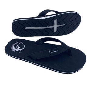 Op de afbeelding: Zwarte slippers met een witte streep rond de rand. De band heeft een wit kruis en de tekst "Johannes 3:16". De zool heeft een wit kruis en de tekst "Loop in zijn voetsporen" in een cirkel.