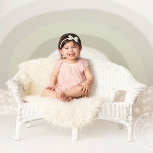 May include: A young child wearing a pink dress and a green and white bow headband sits on a white wicker couch with a white fluffy rug. The child is smiling and looking at the camera.