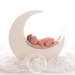 Könnte beinhalten: Ein neugeborenes Baby schläft auf einem weißen, sichelförmigen Mond-Requisit. Das Baby trägt ein weißes Stirnband mit einer Blume. Das Requisit steht auf einer weißen, flauschigen Oberfläche. Das Bild enthält den Text "My Big Camera".