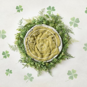 May include: A green knitted baby blanket in a white bowl surrounded by a wreath of green leaves and shamrocks.
