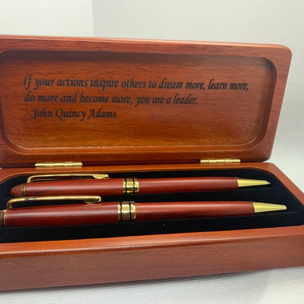 May include: A rectangular wooden box with a hinged lid containing two matching wooden pens with gold-colored clips and tips. The interior lid features the engraved quote: If your actions inspire others to dream more, learn more, do more and become more, you are a leader. - John Quincy Adams.