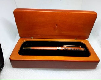 May include: A wooden pen with gold-colored accents and the name Zac Buzza engraved on the barrel, resting inside a rectangular wooden presentation box with a hinged lid and a black velvet-lined interior for secure storage and display.