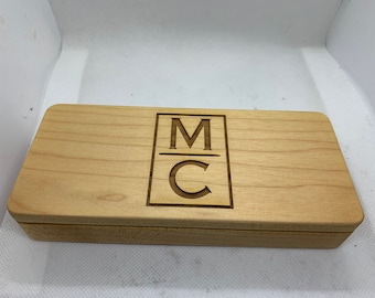 Può includere: Una scatola rettangolare in legno chiaro. Il coperchio presenta un design inciso al laser con le lettere "M" sopra "C" all'interno di un quadrato. La scatola è chiusa e ha una finitura naturale.
