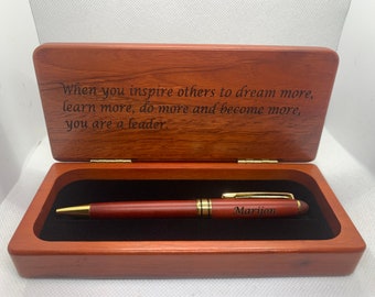 Può includere: Un astuccio per penna in legno con coperchio incernierato che riporta il testo inciso: When you inspire others to dream more, learn more, do more and become more, you are a leader. All'interno, una penna in legno abbinata con dettagli dorati e il nome Marijon.