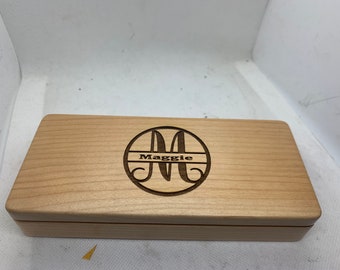 Può includere: Una scatola di legno color chiaro con coperchio incernierato. La parte superiore della scatola presenta un disegno circolare con la lettera "A" e il nome "Maggie" incisi in colore scuro. La scatola è rettangolare e sembra essere fatta di legno naturale.