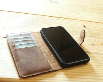 Puede incluir: Una funda de teléfono de cuero marrón con ranuras para tarjetas y una solapa que se pliega sobre el teléfono. El teléfono es negro y tiene una pantalla apagada.