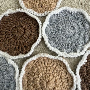 Op de afbeelding: Een verzameling handgemaakte gehaakte onderzetters in verschillende neutrale kleuren: bruin, beige en grijs. Elke onderzetter heeft een witte geschulpte rand. De onderzetters zijn gerangschikt op een neutraal gekleurd, getextureerd oppervlak.
