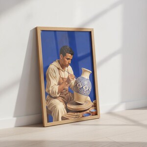 Peut inclure: Une impression d'art encadrée représentant une personne vêtue d'un haut à capuche beige et d'un tablier marron, assise devant un tour de potier. La personne peint un vase en céramique à motifs bleus et blancs sur un fond bleu vif.