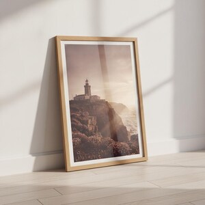 Könnte beinhalten: Ein gerahmter Kunstdruck, der einen Leuchtturm auf einer zerklüfteten Klippe über dem Ozean bei Sonnenuntergang zeigt. Die Szene zeigt felsiges Gelände mit niedriger Vegetation, weiches goldenes Licht und einen dunstigen Himmel, alles in einem hellen Holzrahmen.
