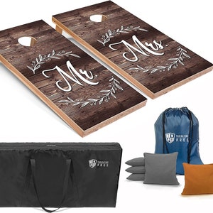 May include: A set of two wooden cornhole boards with a rustic woodgrain design. The boards are decorated with white floral wreaths and the words "Mr." and "Mrs." in a white script font. The set includes a black carrying bag, a blue drawstring bag, and six bean bags: two gray, two orange, and two brown.
