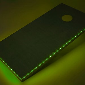 May include: A green wooden cornhole board with a green LED light strip around the edge. The board is angled and the light is illuminating the surface.