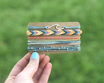 Op de afbeelding: Een set van zes kleurrijke vriendschapsarmbanden met een zilveren golfbedel. De armbanden zijn gerangschikt op een bruine kaart.