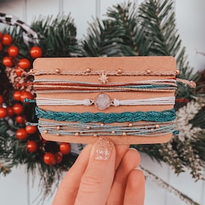May include: A set of eight colourful friendship bracelets with a variety of textures and materials, including braided cord, beads, and a star charm. The bracelets are displayed on a brown card.