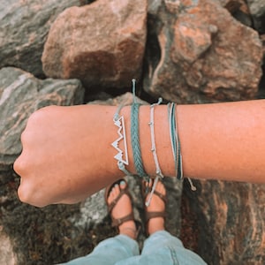 Puede incluir: Una muñeca con una pila de cuatro pulseras. Las pulseras están hechas de cordón fino trenzado en tonos azules y blancos. La pulsera superior presenta un dije plateado con forma de cordillera.