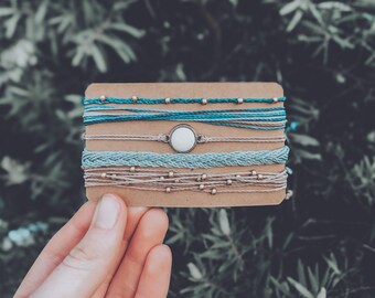 Op de afbeelding: Een set van vijf armbanden met een witte schelpbedel. De armbanden zijn gemaakt van blauwe en beige draad met gouden kralen.