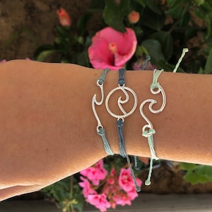 Puede incluir: Tres pulseras de amistad azules y verdes con dijes plateados. Los dijes son un gancho, una ola en un círculo y una ola.