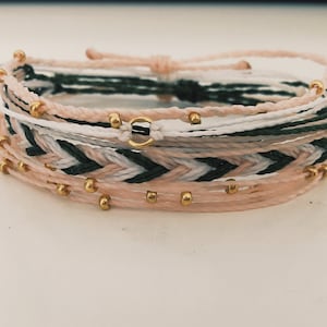 以下が含まれることがあります： ピンク、白、緑の色の組み合わせで編まれた、重ね付けできるブレスレットのセット。ブレスレットには小さな金色のビーズがあしらわれています。