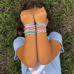 Puede incluir: Una persona lleva una camiseta azul claro con un diseño floral y un parche de lentejuelas. Lleva una pila de pulseras de amistad de colores en ambas muñecas.