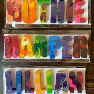 May include: Three crayon name sets: RUTHIE, HARPER, and LILLIAN. Each name is crafted from colorful, swirled crayons, packaged in clear plastic. The crayons are in various colors, including red, orange, green, blue, and purple, on a white background.