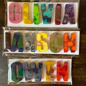 May include: Three sets of handmade crayon names: "OLIVIA", "JAXSON", and "OWEN". Each name is crafted from colorful, swirled wax crayons, packaged on white cards in clear cellophane. The crayons are in various colors, including pink, green, yellow, and orange.