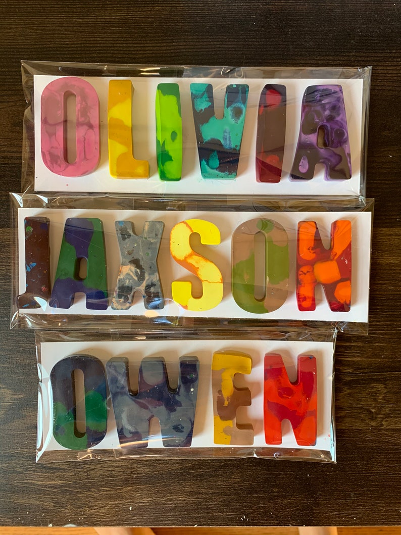 May include: Three sets of handmade crayon names: "OLIVIA", "JAXSON", and "OWEN". Each name is crafted from colorful, swirled wax crayons, packaged on white cards in clear cellophane. The crayons are in various colors, including pink, green, yellow, and orange.
