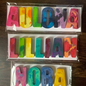 May include: Three sets of colorful, handmade crayon names: "Aulona", "Lillian", and "Nora". Each name is crafted from individual, multi-colored crayon letters, packaged in clear cellophane. The crayons display a variety of hues, including pink, yellow, blue, and green.