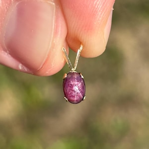 May include: A small oval-shaped cabochon star ruby pendant set in a gold-toned metal prong setting. The deep magenta-colored gemstone displays a distinct six-rayed star pattern across its smooth, polished surface, suspended from a gold-toned bail.