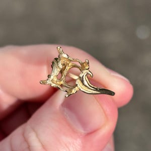 May include: A gold-colored, abstract-shaped earring. The earring has an open, intricate design with curved and pointed elements. The earring is held between fingers, with a blurred background.