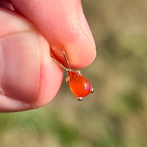 May include: A teardrop-shaped orange fire opal pendant set in a gold metal prong setting. The gemstone exhibits a vibrant, translucent orange hue with subtle flashes of internal color, suspended from a simple gold metal triangular bail.