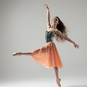 Może przedstawiać: Baletnica w wdzięcznej pozie, ubrana w turkusowy gorset i brzoskwiniową spódnicę. Jej ramiona są wyciągnięte, a ona stoi na palcach w baletkach. Włosy tancerki powiewają, a tło jest miękkie szare.