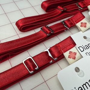 Könnte beinhalten: Mehrere rote Strumpfhalterriemen mit silberfarbenen Metallbeschlägen und kleinen weißen Etiketten mit einem roten Kreuzmuster. Die Riemen sind auf einer weißen Oberfläche mit einem Rastermuster angeordnet.