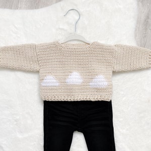 May include: A beige crocheted sweater with white cloud appliques, paired with black jeans. The sweater is hanging on a white hanger.