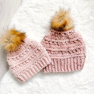 May include: Two pink crocheted hats with brown faux fur pom poms. The hats are on a white fluffy surface.