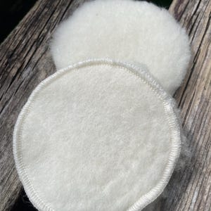 May include: Two white, round, reusable facial cleansing pads. The pads are made of a soft, fluffy material and have a stitched edge. One pad is in focus, while the other is slightly blurred in the background. The pads are designed for skincare.