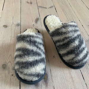 Puede incluir: Un par de zapatillas grises y blancas con rayas de cebra y un forro blanco esponjoso. Las zapatillas están hechas de un material suave y peludo y tienen una suela de goma negra.